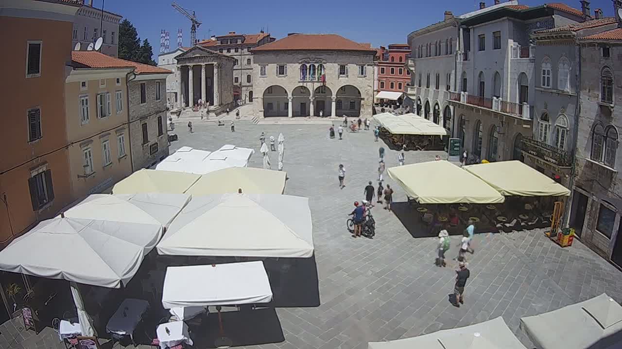A sunny day shows a town square with several outdoor cafes under umbrellas and people walking around historic buildings.