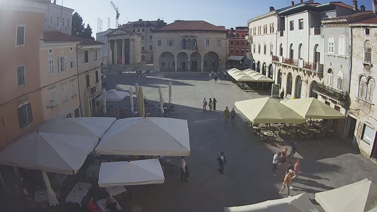 A sunny town square with several outdoor cafes under large umbrellas and a few people walking around.