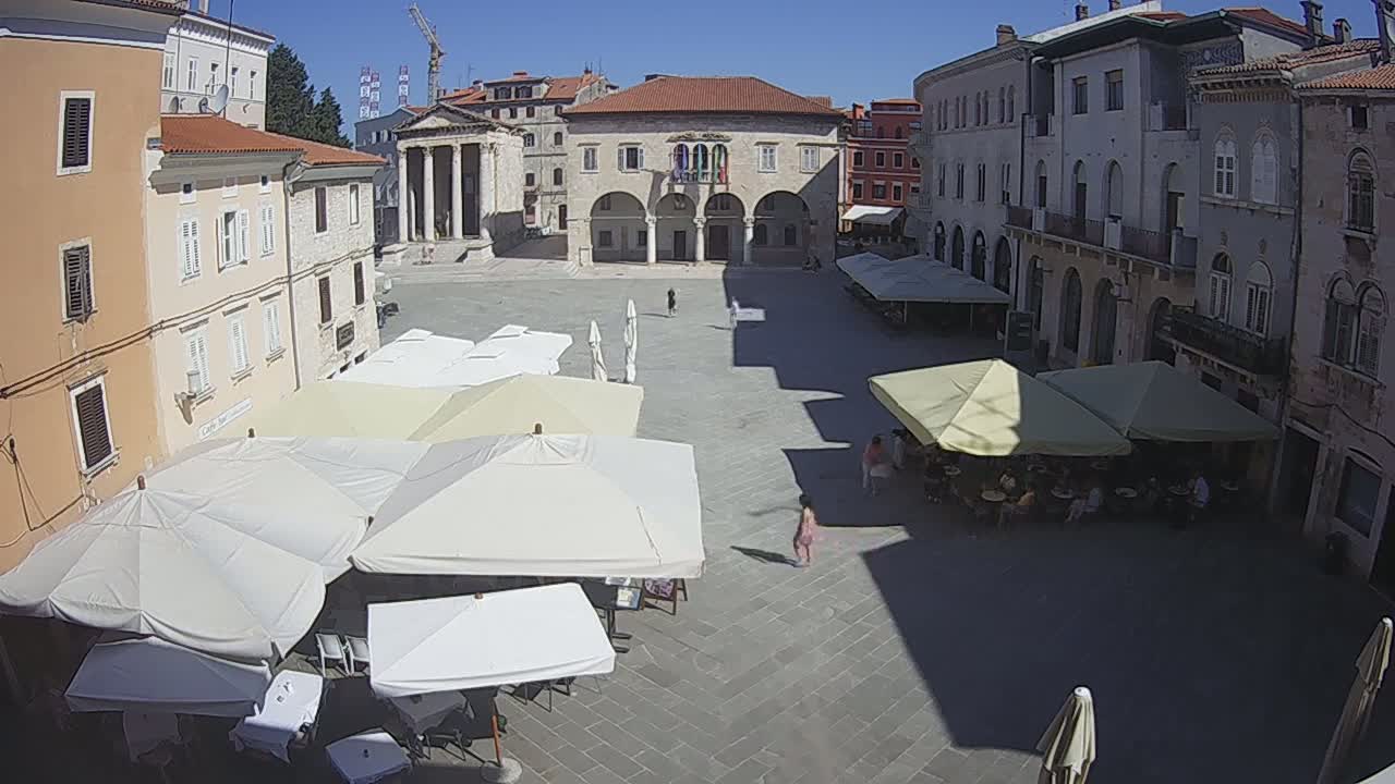A sunny, paved town square is surrounded by buildings, some with outdoor seating under umbrellas.