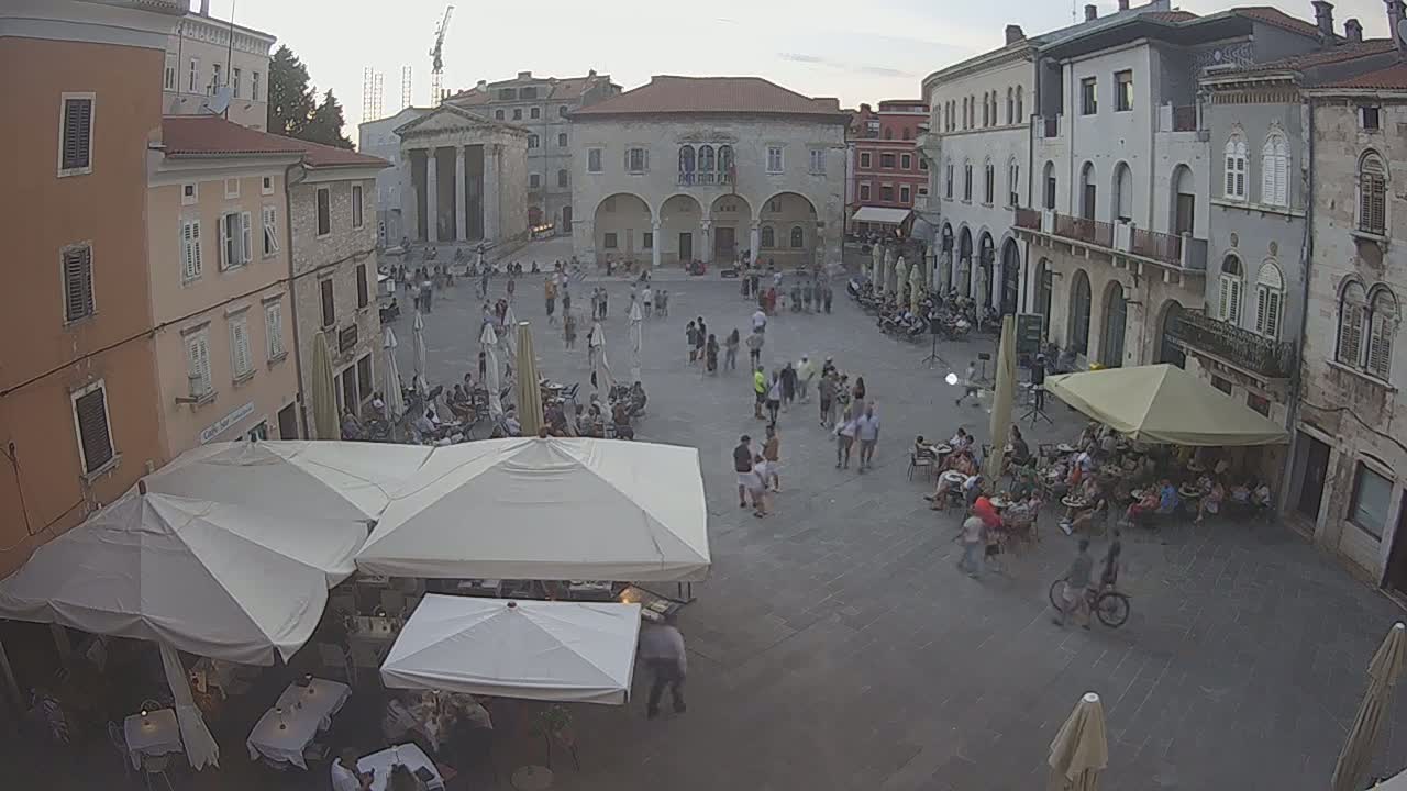 A town square with numerous people milling about, several cafe umbrellas providing shade, and buildings surrounding the paved area under a partly cloudy evening sky.