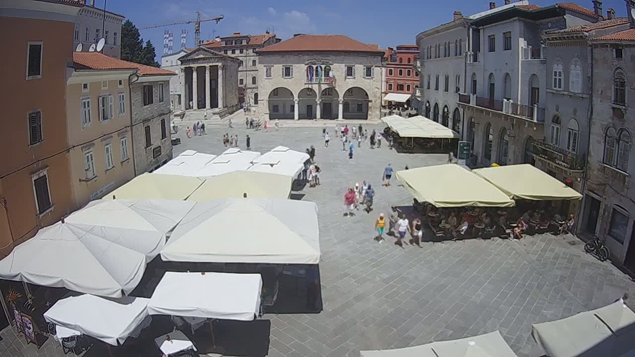 A sunny day reveals a town square filled with people, shaded by numerous outdoor umbrellas and surrounded by historic buildings.