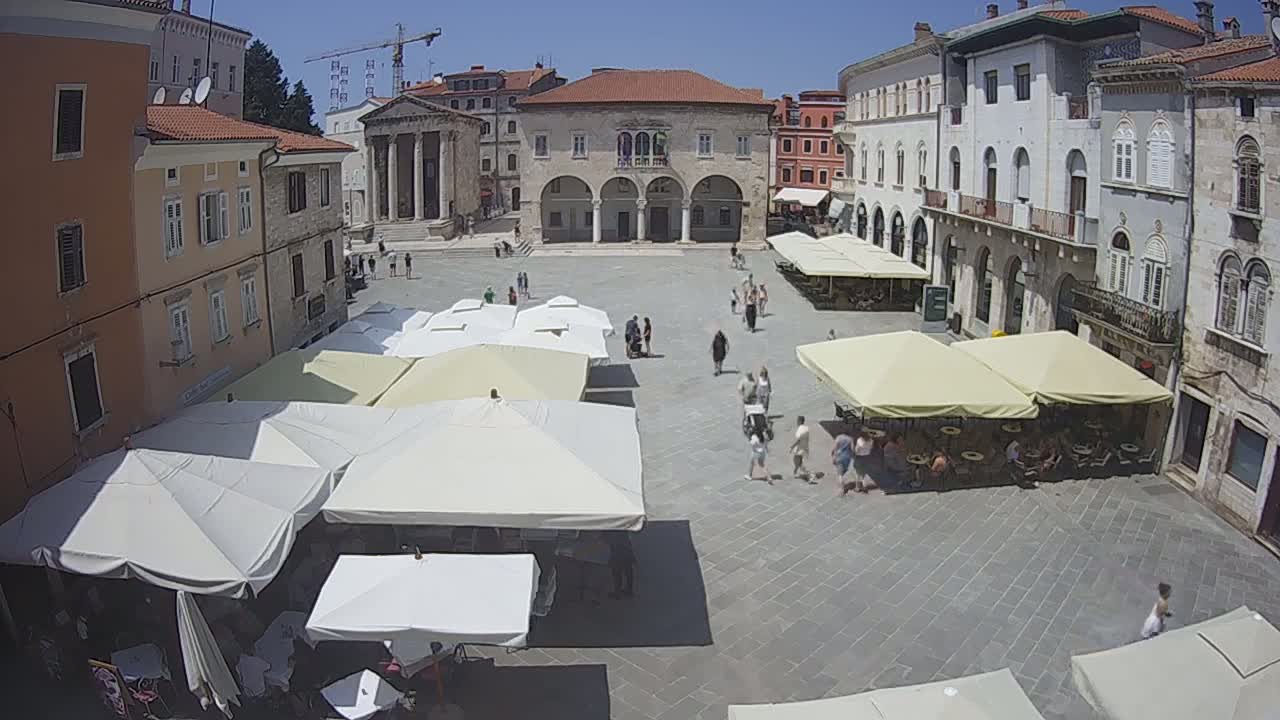 A sunny day reveals a large town square with several people walking around, numerous outdoor cafe umbrellas shading tables and chairs, and historical buildings surrounding the square.