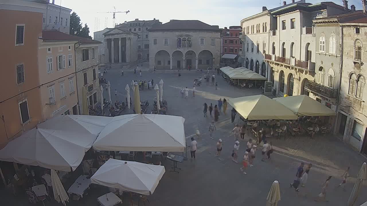 A town square with several outdoor cafes under awnings is filled with people on a sunny day.