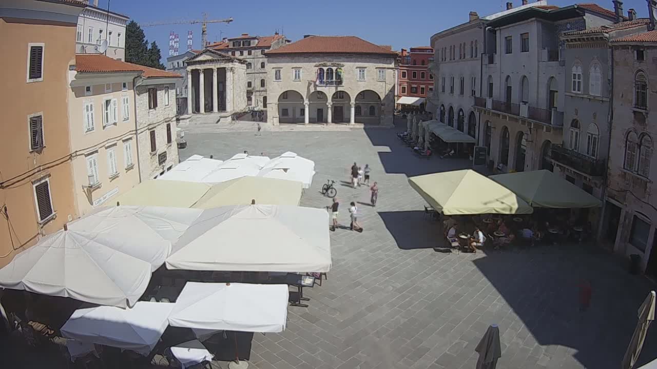 A town square with several people, numerous outdoor café umbrellas, and historic buildings surrounding it on a sunny day.