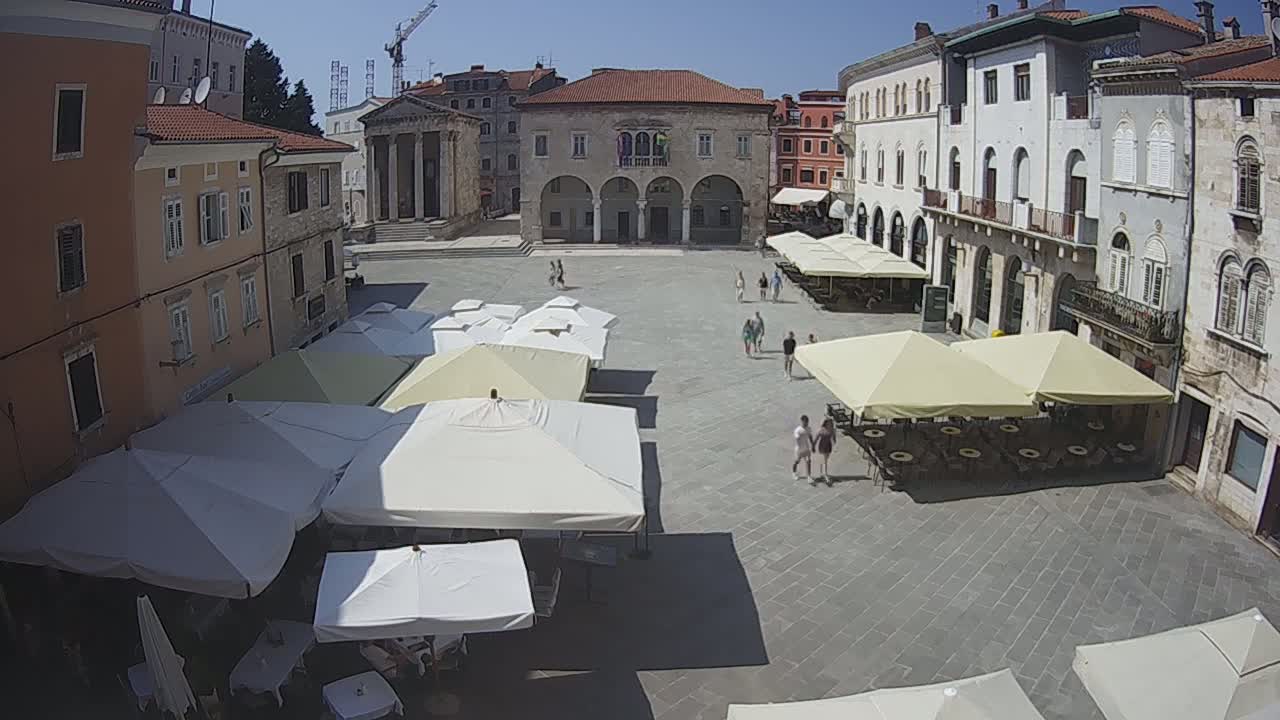 A sunny day in a large, paved town square surrounded by historic buildings, with several outdoor cafe umbrellas and a few people walking around.