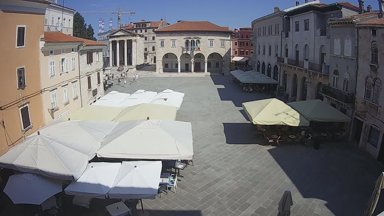 A sun-drenched town square features several outdoor cafe umbrellas under a clear blue sky, surrounding a large building with arches.