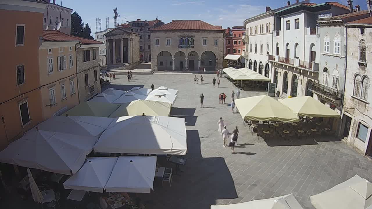 A sun-drenched town square features several outdoor cafe umbrellas under a clear blue sky, surrounding a large building with arches.