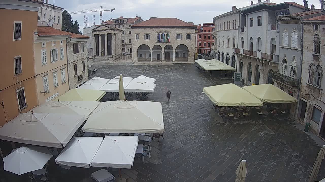 A large, paved town square with several outdoor cafe umbrellas, surrounded by historic stone buildings, under an overcast sky.