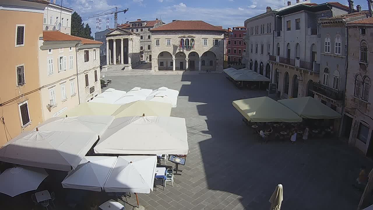 A sunny town square, surrounded by buildings, with numerous outdoor cafe umbrellas and patrons seated at tables.