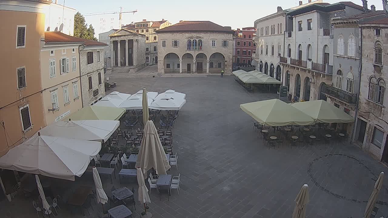 A sunny town square, surrounded by buildings, with numerous outdoor cafe umbrellas and patrons seated at tables.