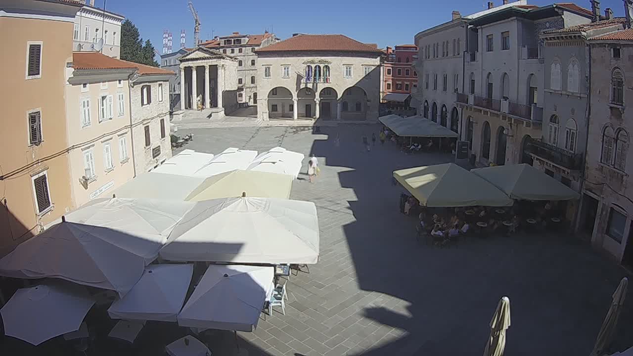 A sunny day in a town square shows several buildings surrounding a paved area with outdoor cafe seating under numerous umbrellas, some patrons are visible.