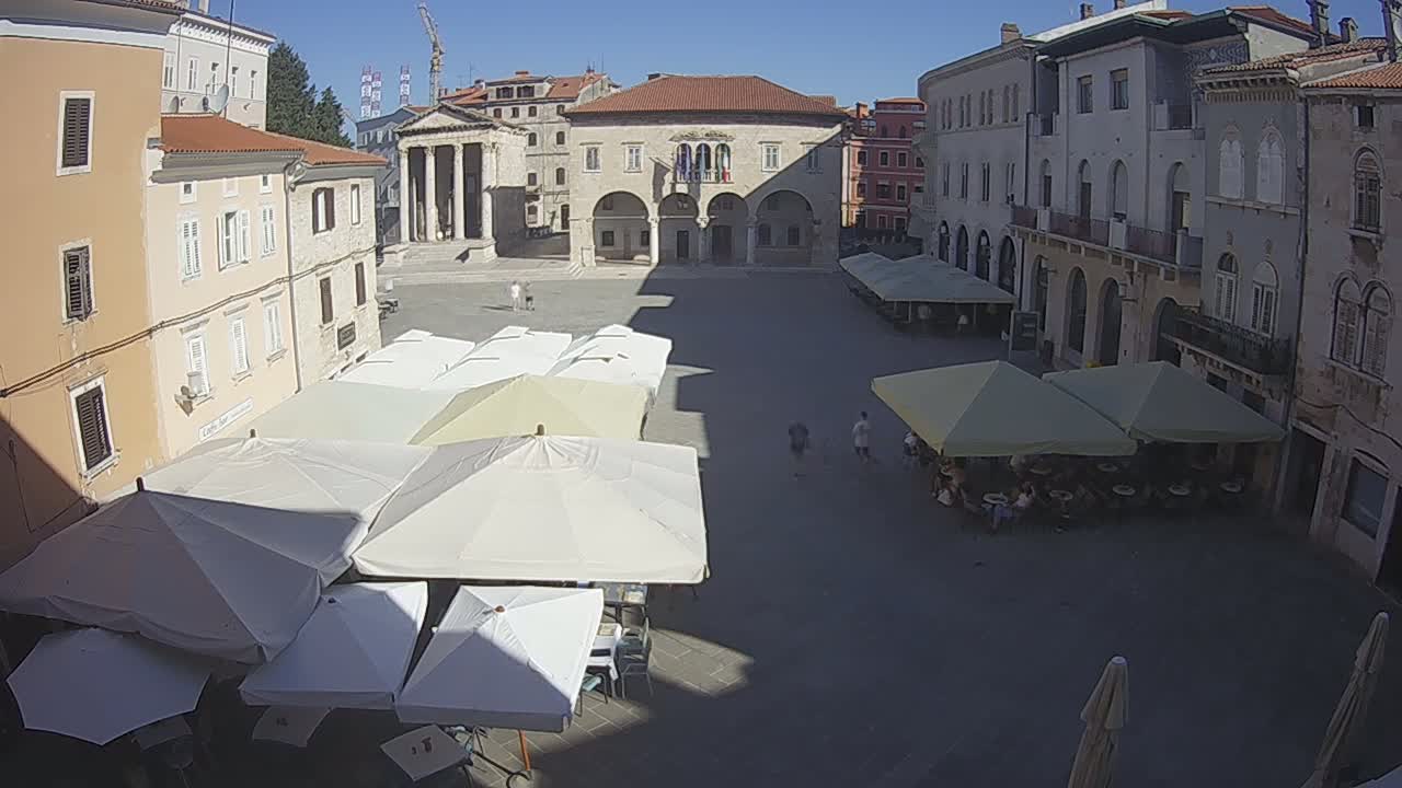 A sunny, outdoor town square with several buildings surrounding it, featuring numerous patio umbrellas shading tables and chairs, and a few people walking.