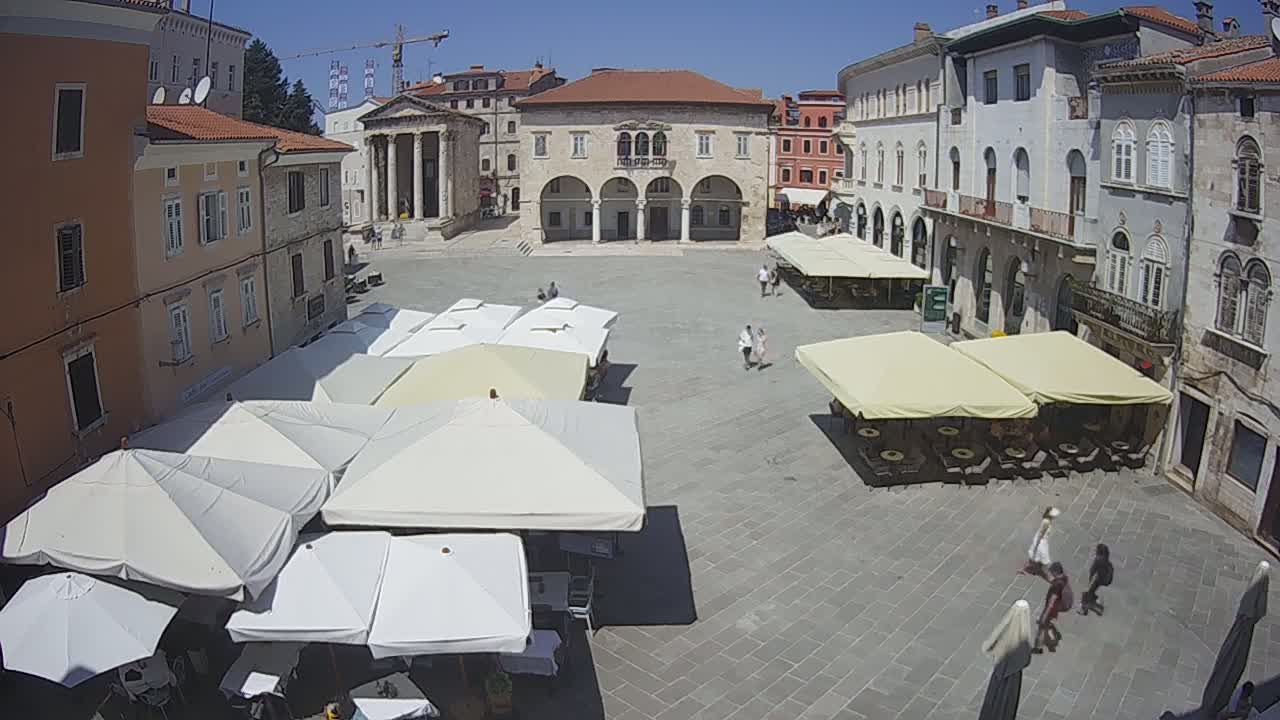 A sunny, outdoor town square with several buildings surrounding it, featuring numerous patio umbrellas shading tables and chairs, and a few people walking.