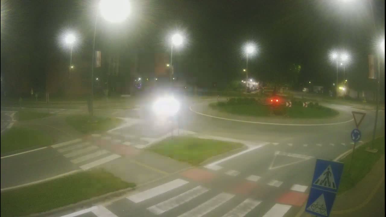 A nighttime view of a roundabout with vehicles and pedestrian crossings, illuminated by streetlights in hazy conditions.