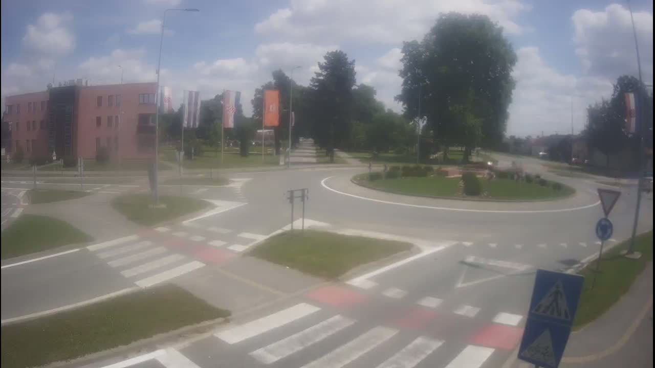 A roundabout intersection on a partly cloudy day, surrounded by buildings, trees, and pedestrian crossings.