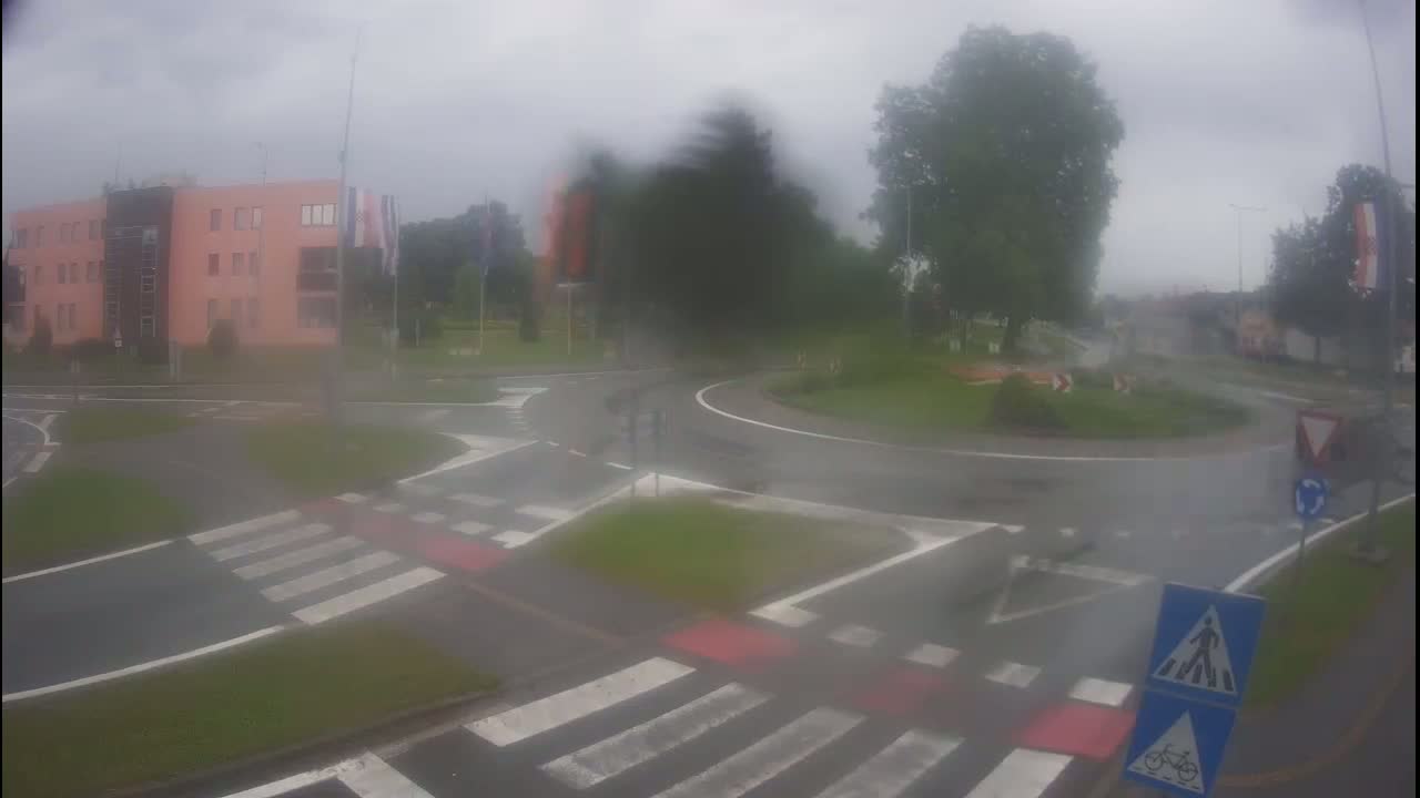 A roundabout in the rain, with a light-pink building and trees in the background.