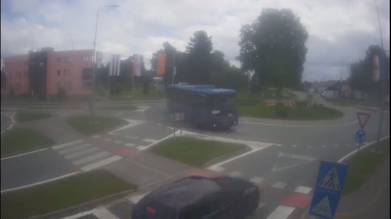 A blue bus navigates a roundabout on an overcast day, with a car partially visible in the foreground.