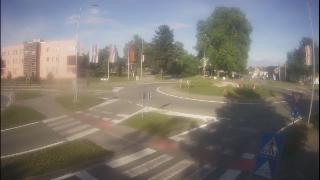 A roundabout surrounded by trees, buildings, and pedestrian crossings under a mostly sunny sky.
