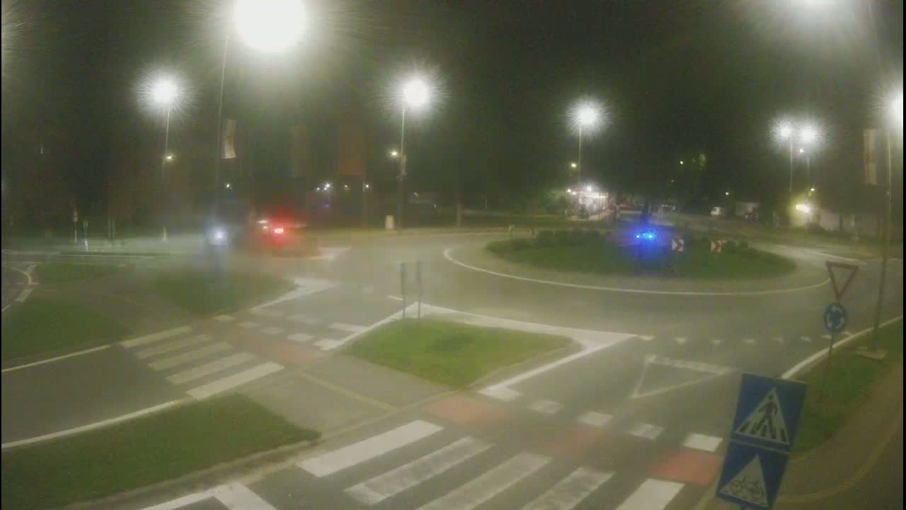 A nighttime view of a roundabout with several cars and pedestrians, under hazy conditions with streetlights illuminating the scene.