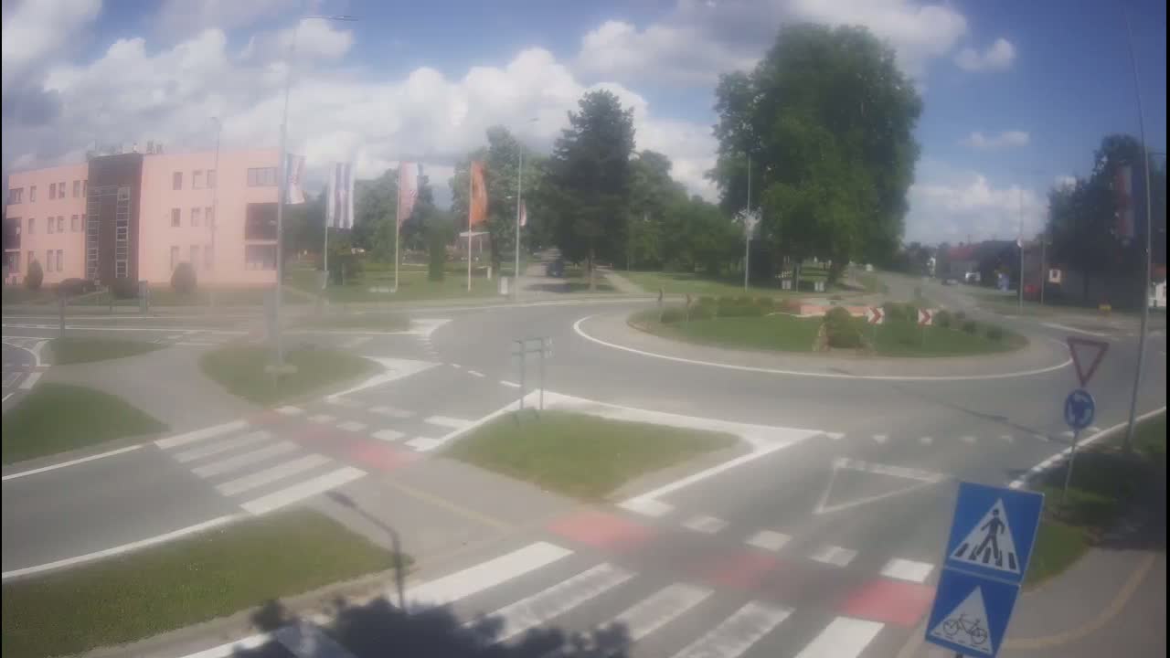 A roundabout with pedestrian crossings and a light pink building in the background under a partly cloudy sky.