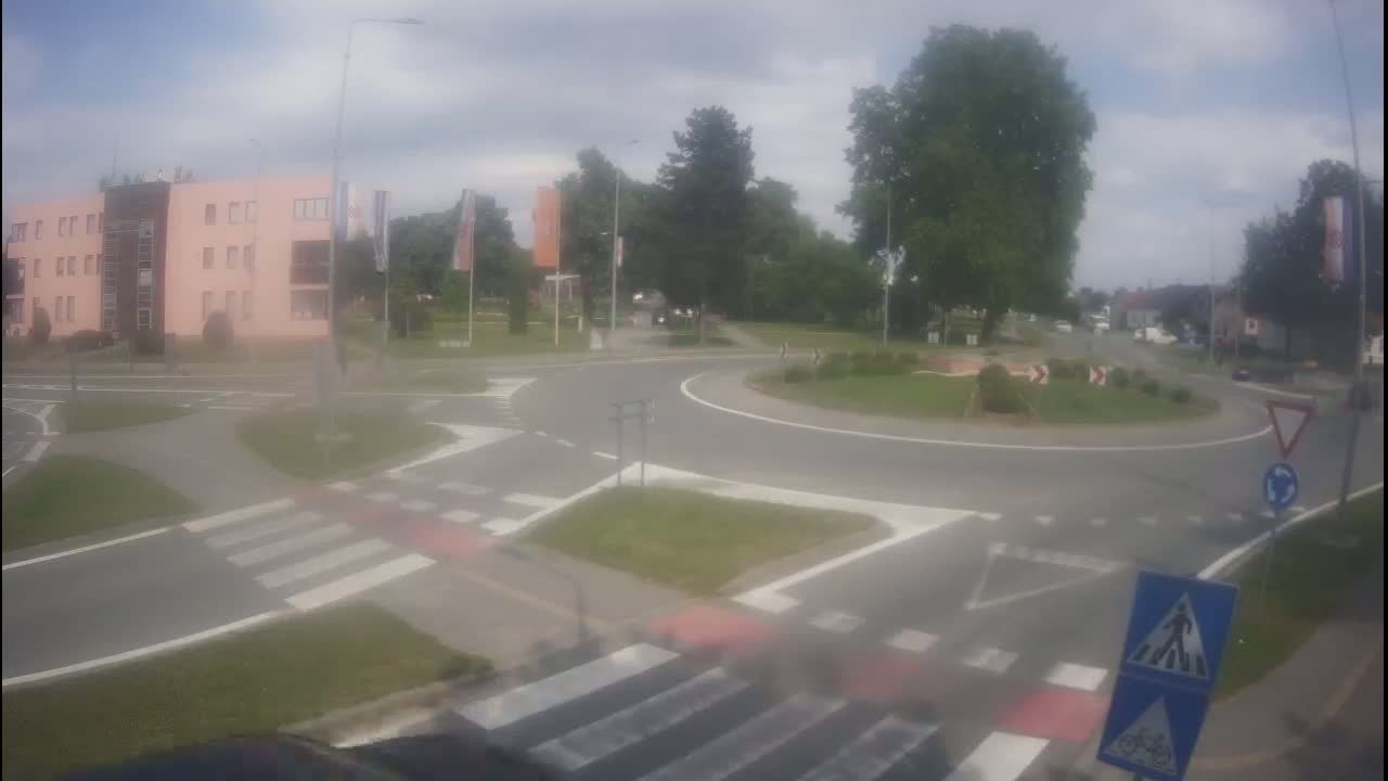 A roundabout with pedestrian crossings is viewed from a car on a partly cloudy day.