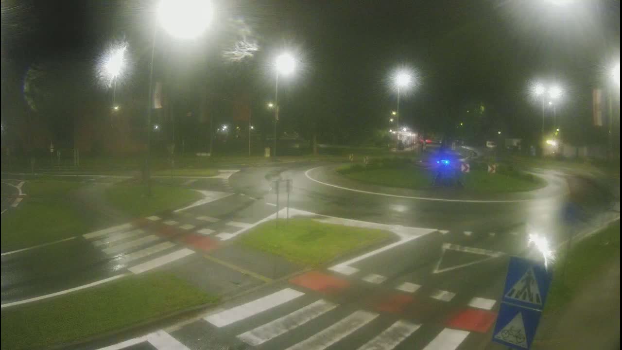 A wet roundabout at night, illuminated by streetlights, with crosswalks and a few vehicles visible.