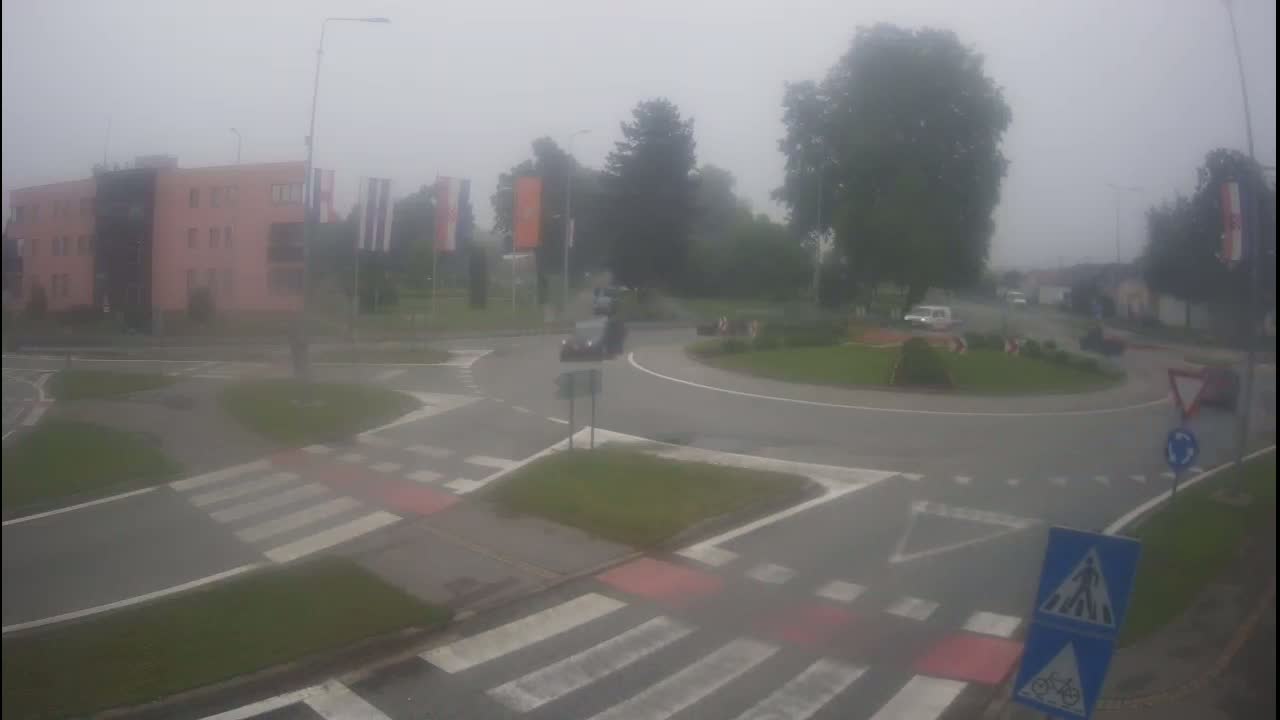A foggy roundabout with several cars, a pedestrian crossing, and a pink building in the background.