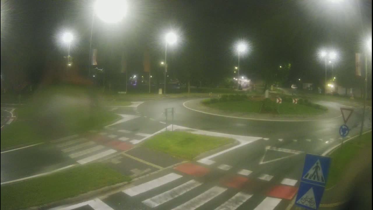 A wet roundabout at night, illuminated by streetlights, with pedestrian crossings and traffic signs visible.