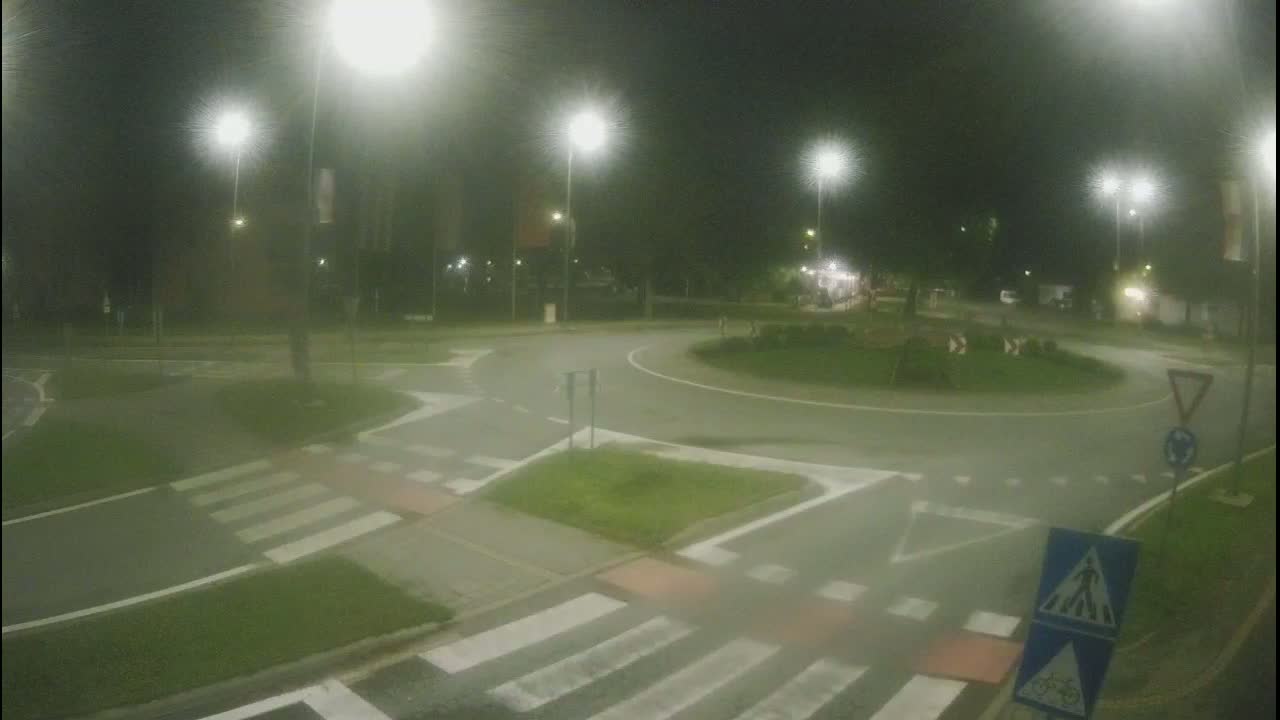 A nighttime view of a roundabout intersection with pedestrian crosswalks, illuminated by streetlights in hazy conditions.