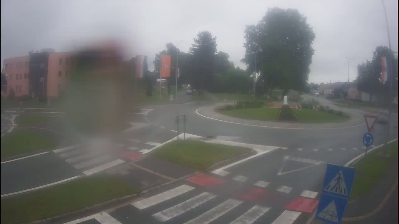 A rain-soaked roundabout with pedestrian crossings is surrounded by buildings and trees.
