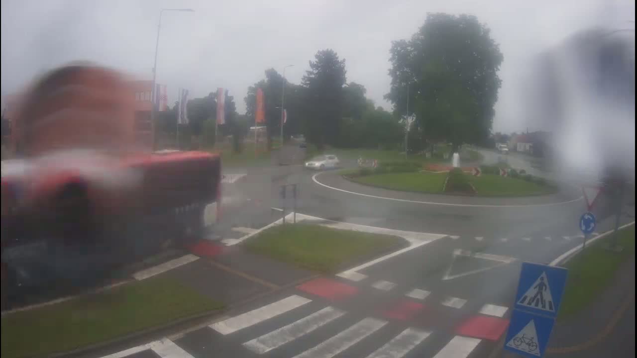 A wet, blurry view shows a car navigating a roundabout, with a bus partially visible to the left, all under a gray, rainy sky.