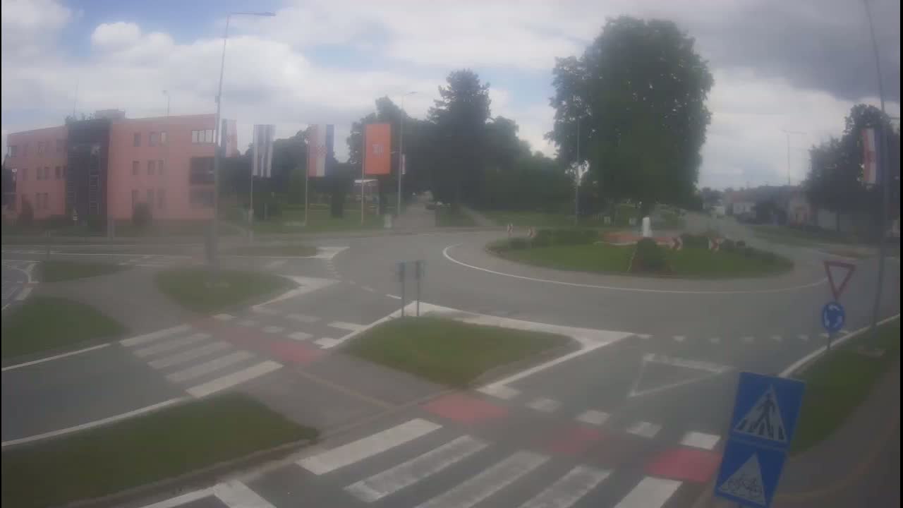 A roundabout with pedestrian crossings is situated near a pink building and lush green trees under a cloudy sky.