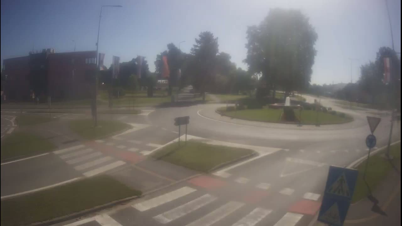 A hazy, sunny day shows a roundabout intersection with crosswalks, a building, and green landscaping.