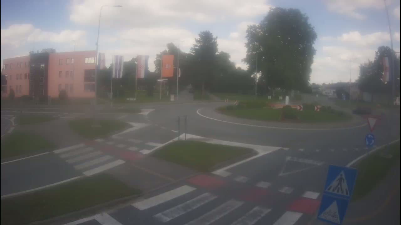 A roundabout with a pedestrian crossing and a light pink building is visible on an overcast day.