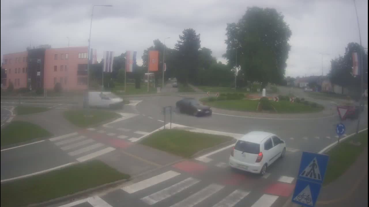 A roundabout with several cars and a light-pink building is visible under an overcast sky.