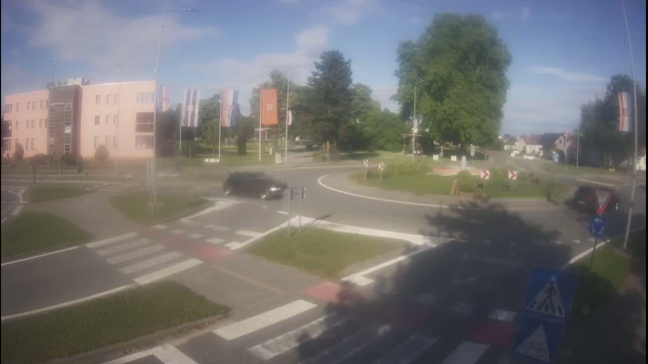 A roundabout with several cars, a light pink building, and lush green trees under a partly cloudy sky.