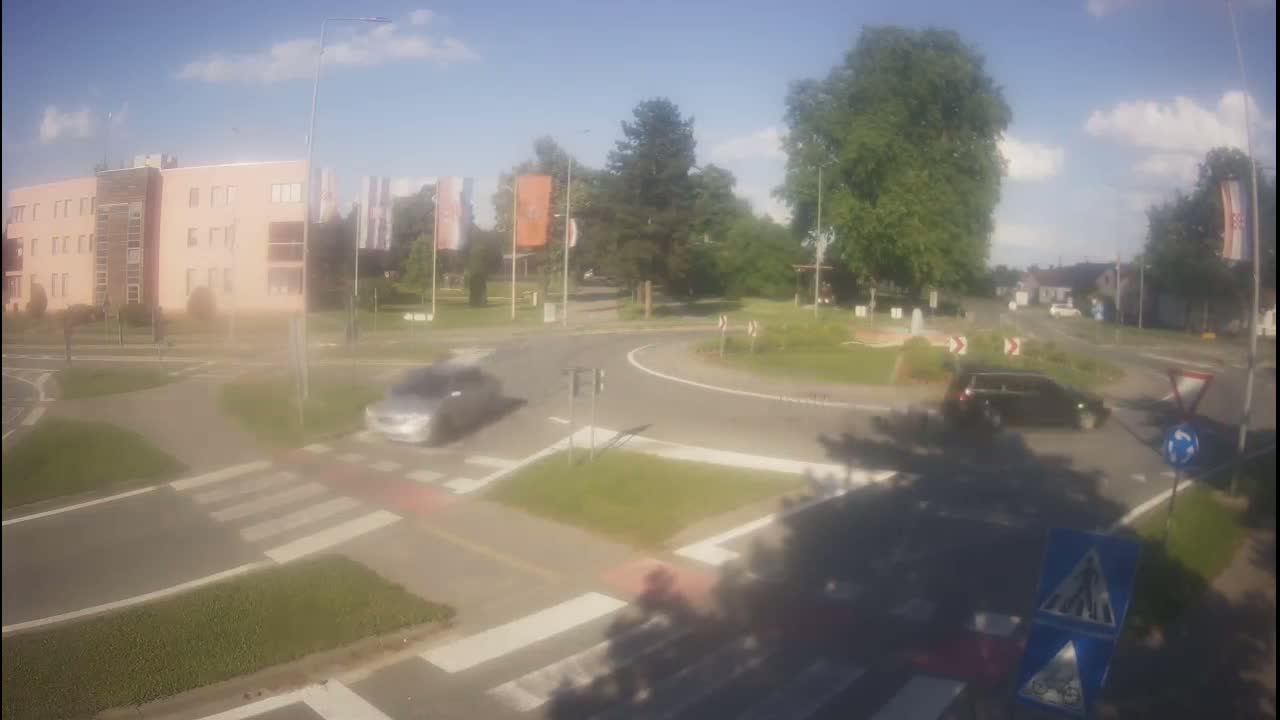 Two cars are traveling through a roundabout on a sunny day, near a light-pink building and lush green trees.