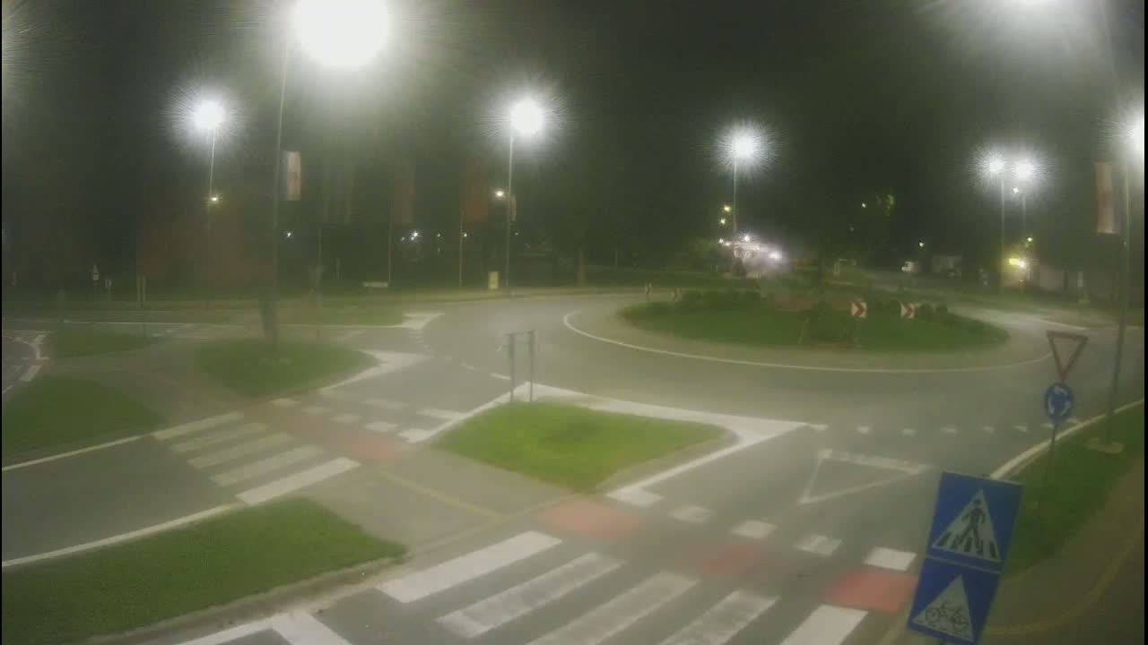 A nighttime view of a roundabout intersection with pedestrian crossings and streetlights illuminated under hazy conditions.