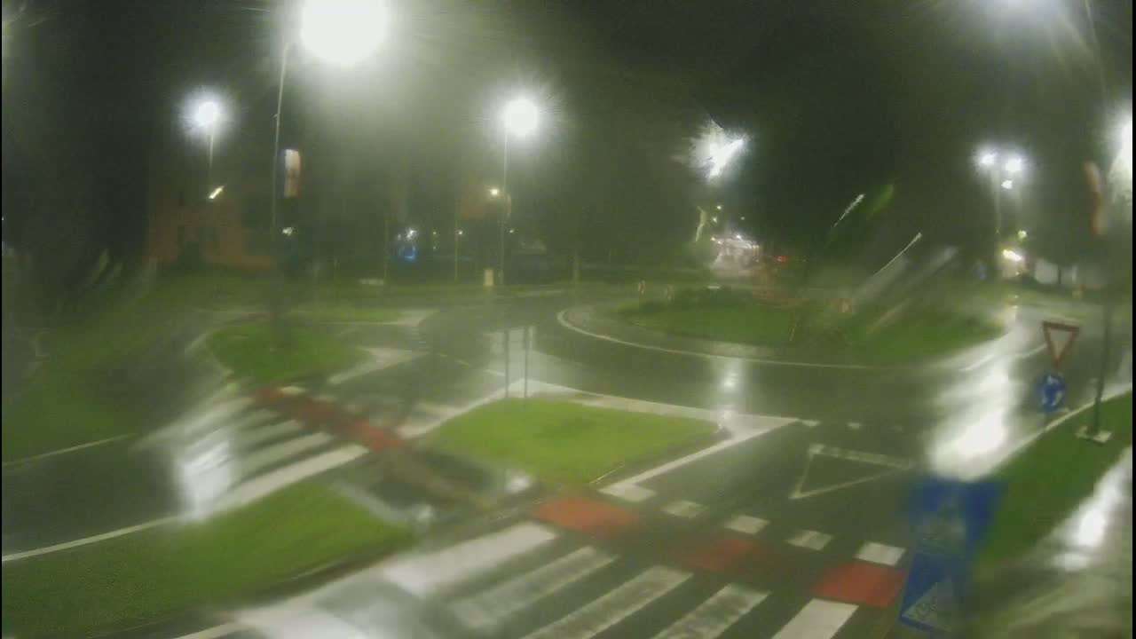 A wet roundabout at night is illuminated by streetlights, with pedestrian crossings visible.