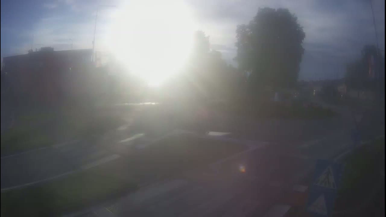 A hazy, bright sun shines over a road and buildings, partially obscured by fog or mist.