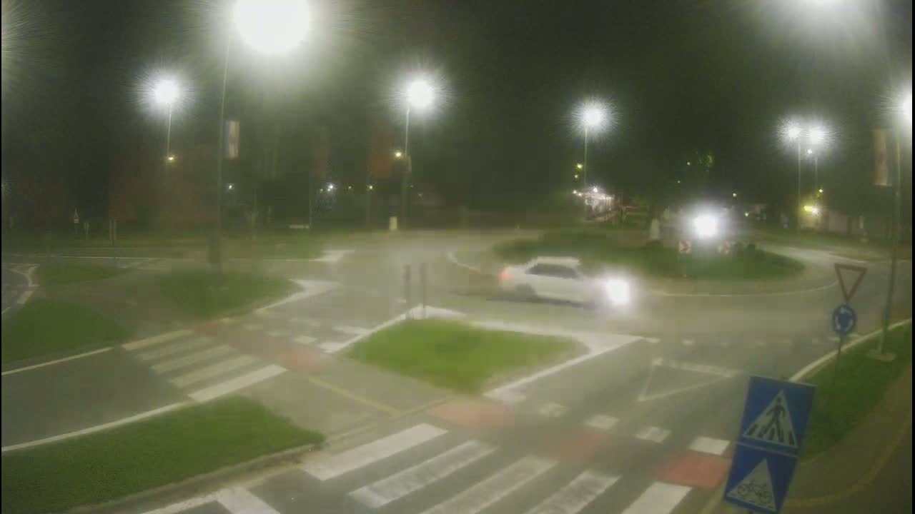 A nighttime view of a wet roundabout with a light-colored car driving through it, surrounded by streetlights and pedestrian crossings.