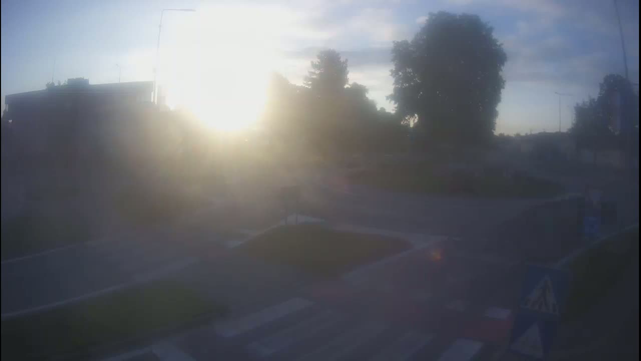 A hazy sunrise illuminates a mostly empty road intersection with several trees and a building in the background.