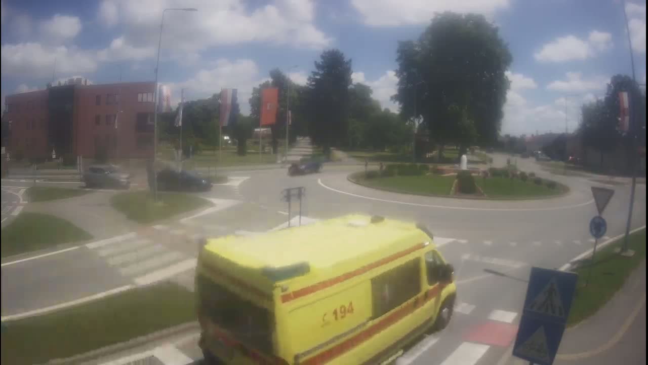 A yellow ambulance approaches a roundabout on a sunny day, with several cars visible around the traffic circle and a brick building in the background.