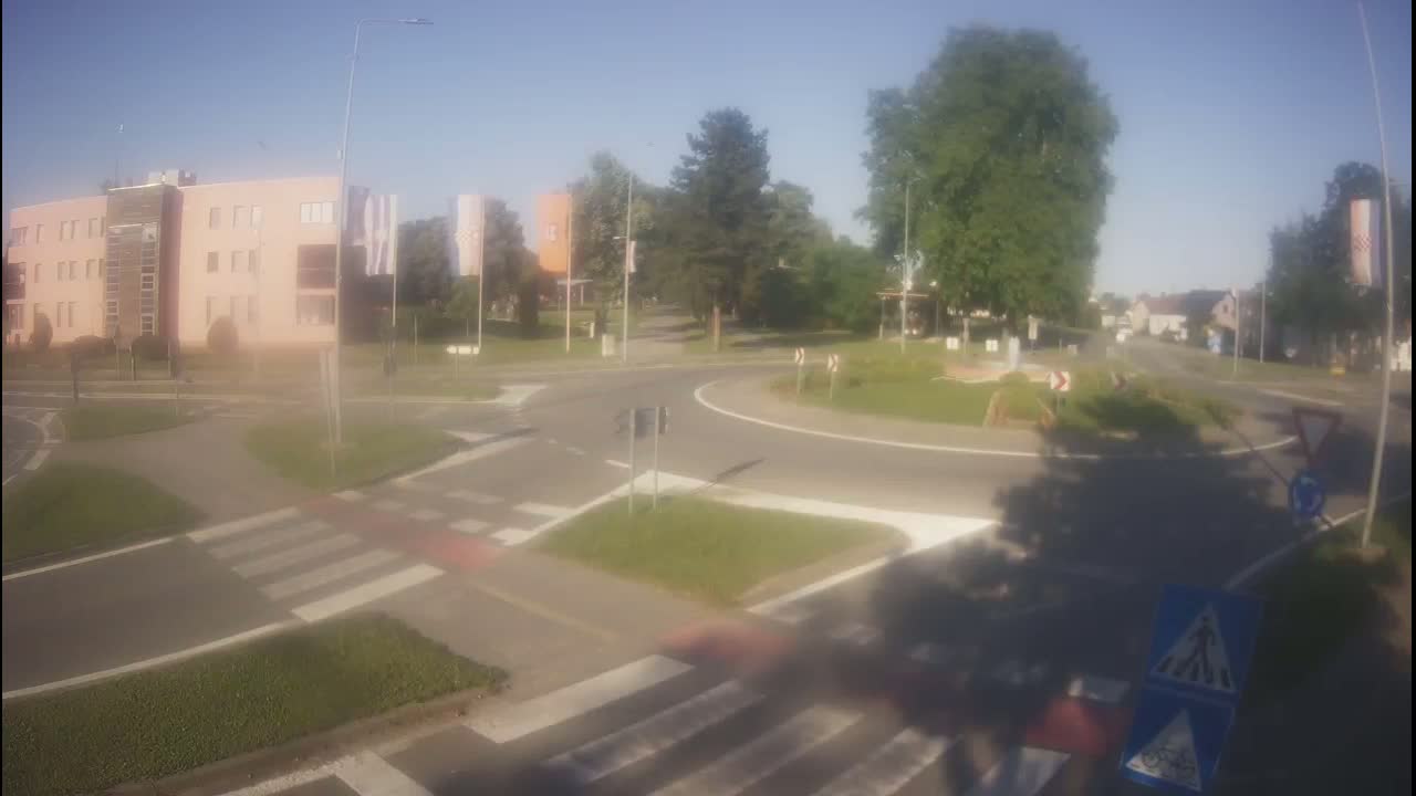 A roundabout intersection on a sunny day, with a light pink multi-story building visible in the background, surrounded by trees and other buildings.