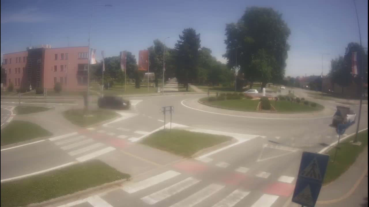 A roundabout with a few cars is surrounded by trees and a pinkish-tan building under a clear, sunny sky.