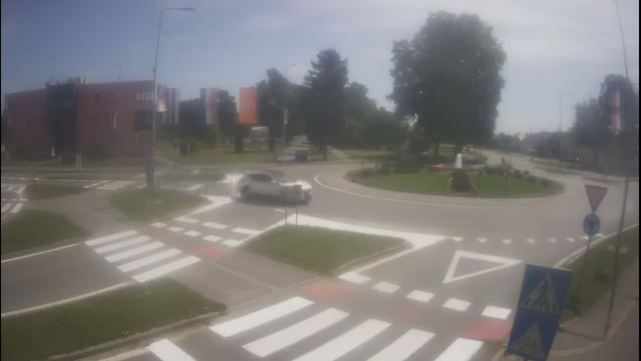 A white car navigates a roundabout on a sunny day, with pedestrian crossings and surrounding greenery visible.