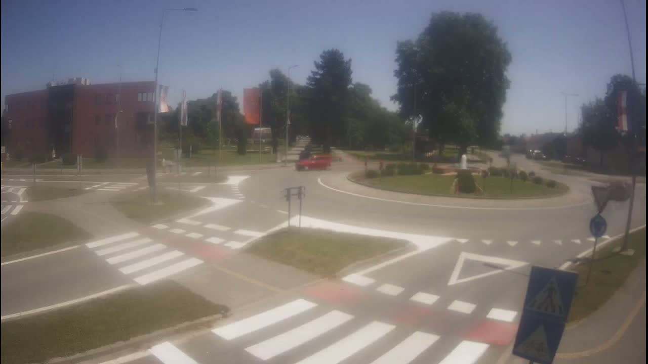 A red car is driving through a roundabout on a sunny day surrounded by buildings and trees.