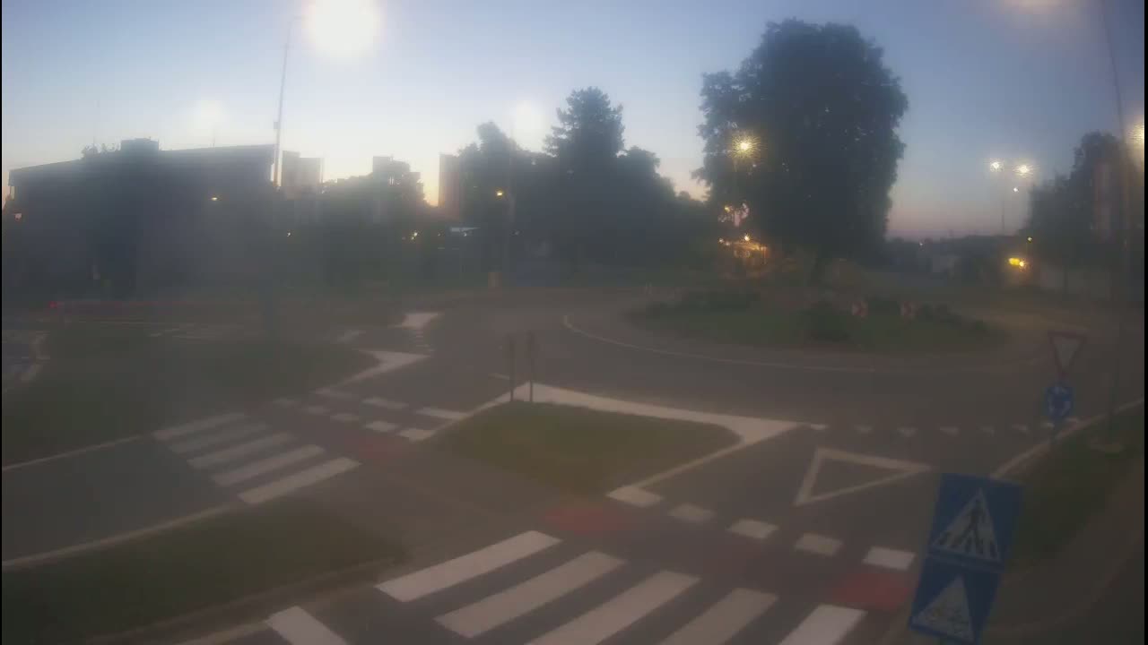 A hazy, twilight view of a roundabout with pedestrian crossings, surrounded by buildings and trees.