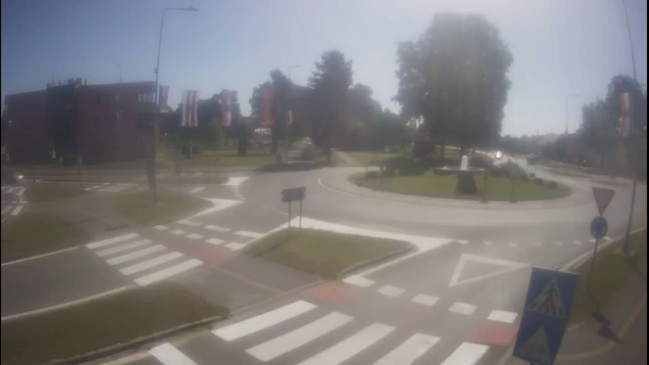 A hazy, sunny day shows a roundabout intersection with pedestrian crosswalks and a few vehicles, surrounded by buildings and trees.