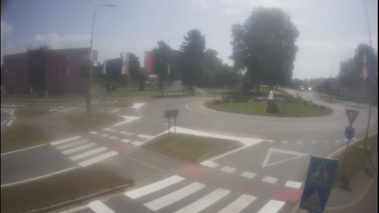 A hazy, sunny day reveals a roundabout intersection with pedestrian crosswalks, surrounded by buildings and green trees.